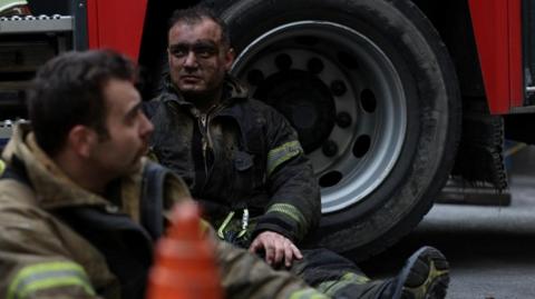 A firefighter sits on the ground, leaning back against the wheel of a fire engine. His face is blackened and he looks off into the distance,