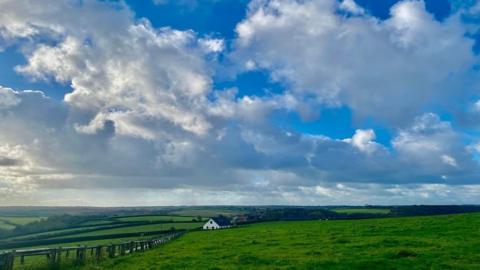 Huge white clouds sit in a blue sky over bright green fields with a white house just to the left of centre.