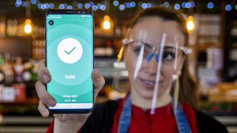 A woman working in a bar holds a phone displaying an approved Covid certificate