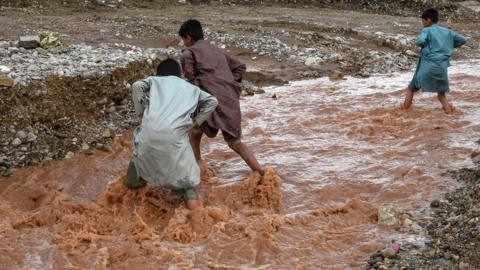 Children wade through floodwater near a damaged road following heavy rains on the outskirts of Quetta on 15 April 2024