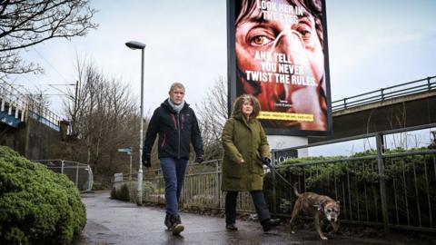 Members of the public walk past a government poster reminding people to socially distance and abide by the lockdown restrictions in the city centre on January 29, 2021