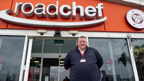 Matt Embleton - with short dark hair wearing a lanyard outside the main entrance of Watford Gap service station