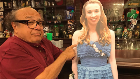 Danny DeVito stands next to a cardboard cutout of a teenager in a prom dress. In the background is the set of Paddy's Pub, where TV show It's Always Sunny in Philadelphia is filmed.