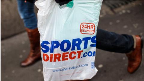 A plastic Sports Direct bag is carried by a shopper.