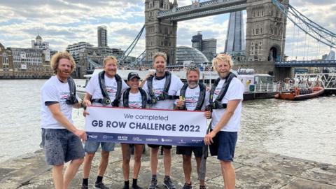 The team after completing the trip under Tower Bridge hold a sign saying "We completed GB Row challenge 2022"