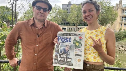 Two people standing on the banks of the River Avon holding an old copy of the Bristol Post