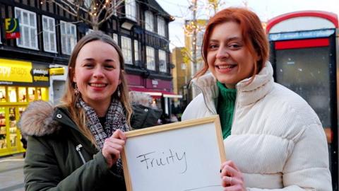 Two women holding a whiteboard with the words "Fruity" and "Adventurous"