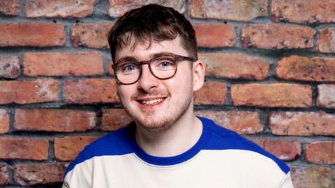 Jack Carroll, announced as joining the Coronation Street cast as Bobby. Jack, a white man in his 20s, has short brown hair and wears tortoiseshell rimmed glasses. He has blue eyes and a short stubble and smiles at the camera. He is pictured in front of a brick wall wearing a blue and white striped top.