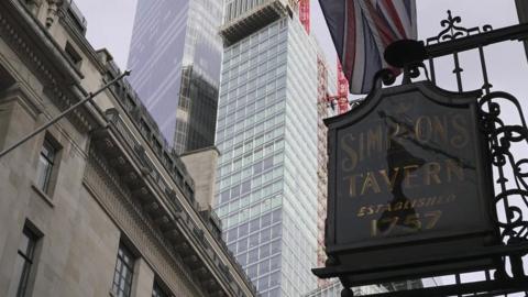 An ornate green and gold sign for Simpson's hangs in front of a backdrop of skyscrapers in the city of London.