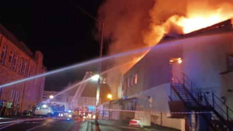 Firefighters stand in street spraying water at building on fire