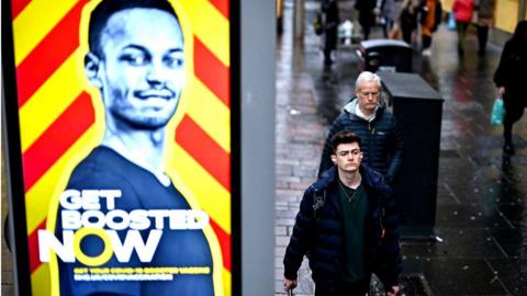 vaccine booster poster in the street with people walking by