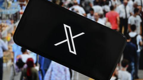'X' (formerly known as Twitter) logo is being displayed on a mobile phone screen in front of a computer screen displaying a photo of a crowded sidewalk,