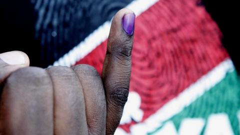 Voter shows finger inked with purple with Kenyan flag colours in background