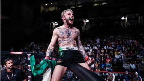 James Gallagher shouts celebrating on the cage wall with an Irish flag