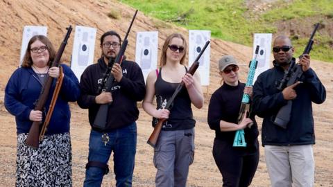 Five people from minority groups holding rifles, standing in front of targets at a shooting range in San Diego.