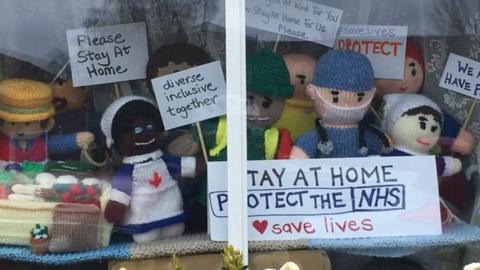 Knitted figures in window with signs "Stay at home. Protect the NHS" and "We stayed at work for you now stay at home for us please"