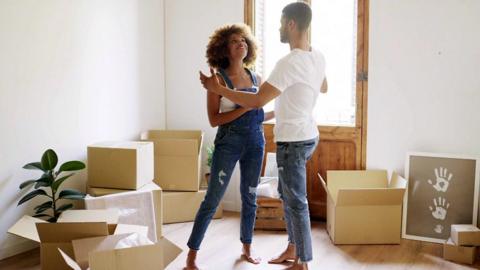 A man and woman stand in a room with cardboard boxes, a house plant and some artwork