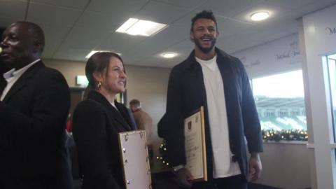 Woman with long brown hair wearing black jacket and tall man with dark hair and beard wearing grey jacket hold framed certificates