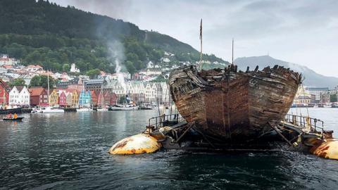 The hull of the Maud is seen in the water in front of a Nordic coastal town