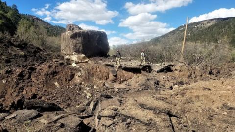 The section of the road that was destroyed by a rock slide containing two enormous boulders with two workers standing in the road.