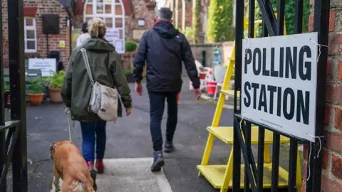 A couple and a dog walk through a gate with the sign 'polling station' in Yarm on 2 May