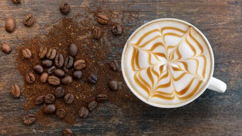 A cappuccino with a caramel web motif pictured next to a pile of coffee beans and grounds.