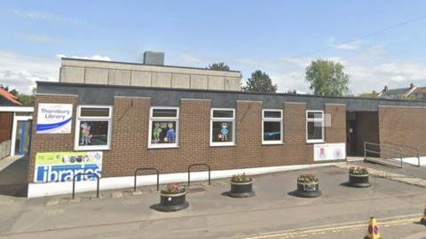 Google Maps view of Thornbury Library. It is a long, red brick building with five windows on it. On the left hand side of the building, there is a white sign reading 'Thornbury Library'