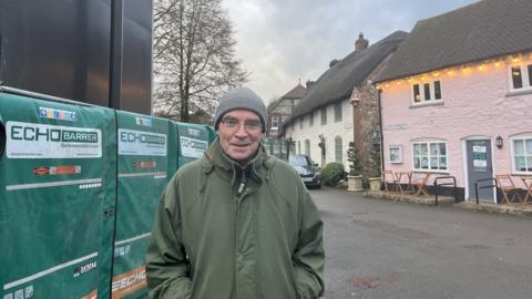 Nick Josephy looks into the camera dressed in a green winter coat, glasses and woolly grey hat. A set of construction fencing and scaffolding to the left, old quaint cottages to the right