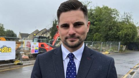 Councillor Joe Harris. He has dark hair and facial hair. He is wearing a navy blue suit jacket, light blue shirt and blue tie with white spots. He is squinting, but looking straight at the camera, with a slight smile.