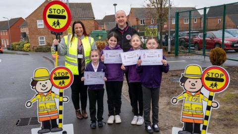 Children along with a teacher and Northamptonshire's Police, Fire and Crime Commissioner, pose outside the school with the signs