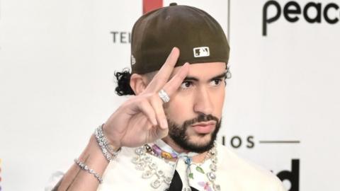 Bad Bunny attending the 2023 Billboard Latin Music Awards in October 2023. Bad Bunny is a Puerto Rican man in his late 20s. He has a short dark beard and wears a brown cap twisted back on his head. He wears a white shirt with a diamond necklace and a neck scarf. He holds his right hand up to his face making a peace sign.