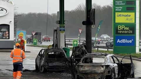 The burnt out shells of two cars on a petrol forecourt.