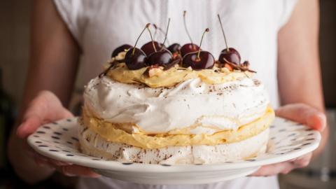 A pavlova with cherries on top is seen on a plate held by a woman