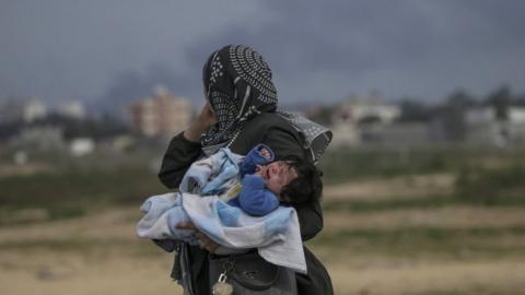 A Palestinian woman carries her baby as smoke rises below her in the Gaza Strip. Photo: 26 February 2024