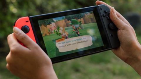 A pair of hands holds a Nintendo Switch console in an outdoor - garden - environment. On the screen is a shot from Animal Crossing - two characters are in conversation, as denoted by an on-screen dialogue box.