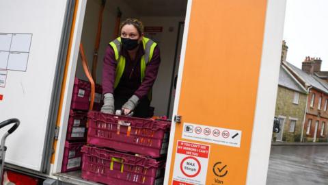 A woman in high-vis and a black mask leans over a crate.