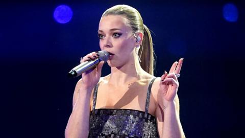 A female singer holds a microphone close to her mouth with one hand, her other hand is held up at her side, two fingers extended and pointing upwards. She's wearing a strappy, sparkly black top. Her hair is tied back in a tight, sleek pony tail and and she's wearing an earpiece.