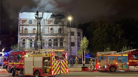 Burning Grosvenor Hotel and fire engines in Bristol. Although the image was taken at night, smoke can be seen rising from the building. Two fire engines are in the foreground of the shot, with a number of crew members stood nearby.