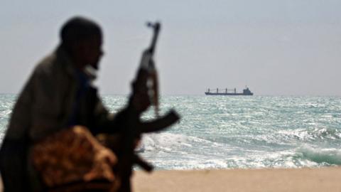 Photo made on January 7, 2010 shows an armed Somali pirate along the coastline while the Greek cargo ship, MV Filitsa, is seen anchored just off the shores of Hobyo town in northeastern Somalia where its being held by pirates