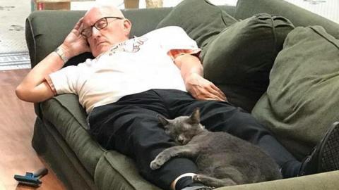 Terry lies on a sofa with a grey cat curled around his legs