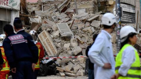 Rescue workers and police stand either side of frame, at the centre of which is a jumble of jumble and loose wooden fixtures where a building once stood.