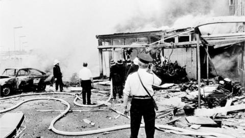 A dark mushroom cloud of smoke drifts across the centre of Belfast, as firemen hose down the remains of Oxford Street bus station.