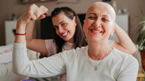 two women, one who has lost her hair, pose for the camera in strong poses with big smiles