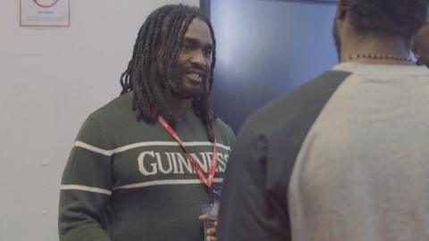 A man with dreadlocks wearing a green jumper with Guinness written across the chest smiles as he listens intently to a man in a grey sweater with his back to the viewer.