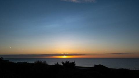 The sun sets over the sea. The sky above is light blue, turning to deeper blue