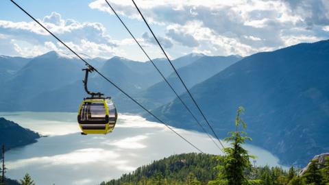 A shot shows a cable car move down towards a stunning lake at the base of rolling green mountains carpeted thickly with pine trees