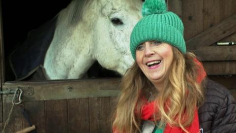 Eva Hamilton. She is stood in front of a white horse. She is looking to the side of the camera and smiling. She is wearing a green bobble hat and a red scarf.