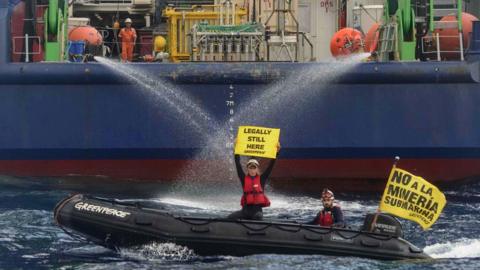 Greenpeace protesters on boat holding banners in front of larger boat
