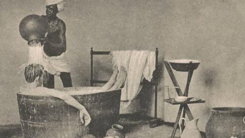 A postcard showing Indian domestic help pouring water over a white man while he bathes in a tub.