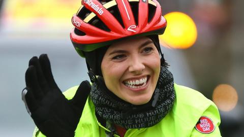 A woman rides a bicycle on a rain-covered road with an escort car trailling behind. She's smiling and waving, and wears a bright red crash helmet and a bright yellow hi-vis jacket.